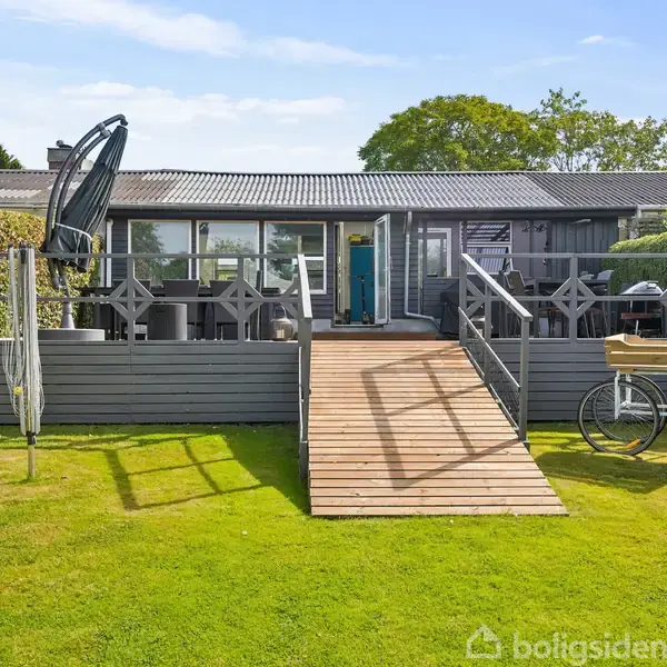 Græsplæne strækker sig mod en rækkehusbebyggelse, hvor en terrasse med trapper fører op til husets bagindgang. Omgivet af hække og træer under en blå himmel.