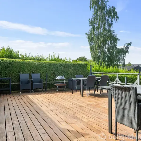 En udendørs terrasse er indrettet med kurvemøbler og en grill, beliggende i en have omgivet af grønne hække og træer under en blå himmel.