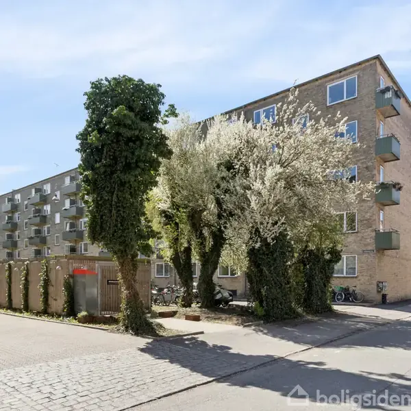 Et beboelsesbyggeri med flere etager står langs en vej. Ved siden af bygningen er der træer og parkerede biler. Sollys oplyser omgivelserne, og der er en blå himmel.