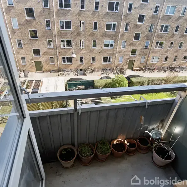 En lille altan med flere urtepotter står stille. Den omgivende kontekst er en udsigt over en gårdhave med cykler og en stor murstensbygning i baggrunden.