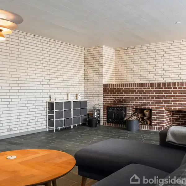Stue med sort sofa, rundt træbord og stor murstenspejs. Væggene er af lyse mursten. Over bordet hænger en moderne lampe.