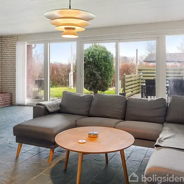 En grå sofa står i en stue med glasdøre, der åbner mod en have. Rummet har hvidt loft, stengulv og en murstensvæg med billeder og en pejs.