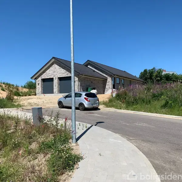 En grå bil står parkeret foran et moderne, gråstenshus med sort tag. Huset har dobbelte garager. Omgivet af græs og buske på en solrig dag.