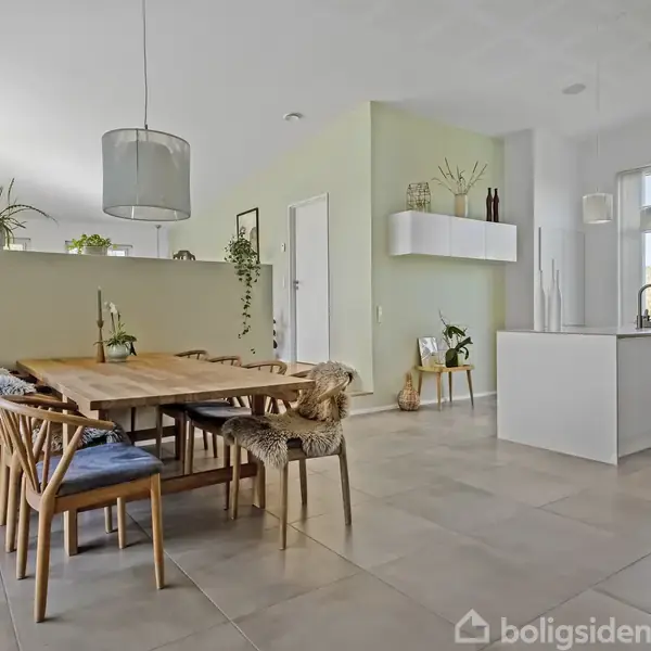 Et spisebord er omgivet af stole i en moderne, lys stue med planter og en køkkenø i baggrunden. Store vinduer giver naturligt lys, og rummets vægge er lysegrønne.