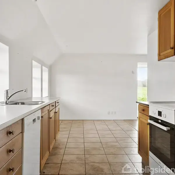 Køkken med træskabe og hvide bordplader, opvaskemaskine og ovn, står i et rum med flisegulv og store vinduer, der skaber en lys atmosfære.