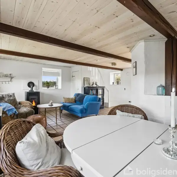 Rundt bord med stole står i stuen. Pejs ved væg, blå sofa ved vindue. Trægulv med tæppe, loftsbjælker. Lys og hyggelig atmosfære i moderne indretning. Markerer skandinavisk stil.