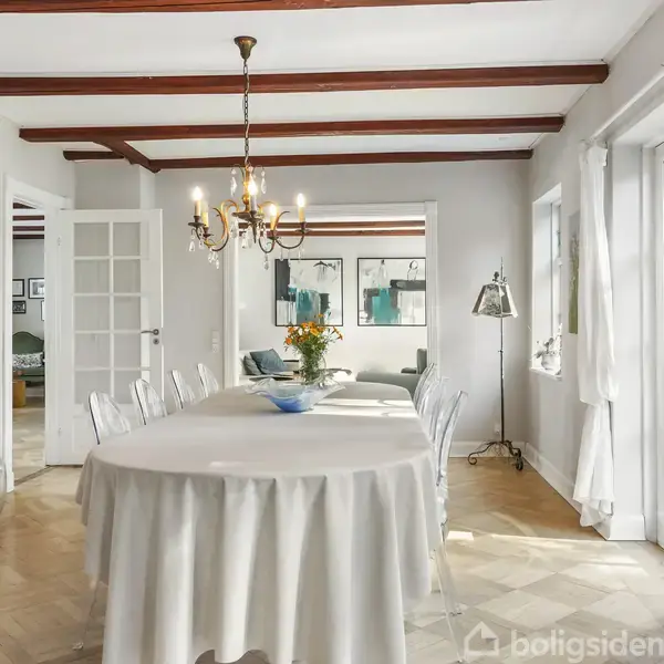 Spisebord dækket med dug står i centrum af en lys spisestue. Over bordet hænger en lysekrone. Der er vinduer og en dør til højre og skænk til venstre.