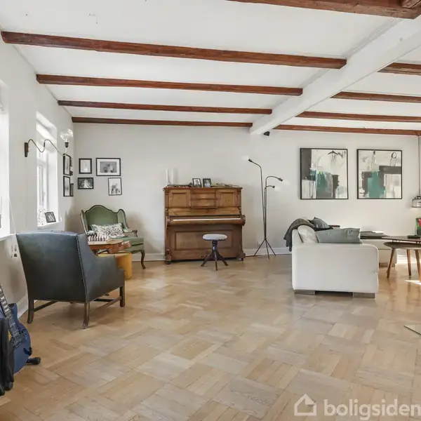 En lys stue med trægulv har en klaver i midten til højre og en guitar i etuiet til venstre. Rummet indeholder også en sofa, stol og flere kunstværker på væggene.