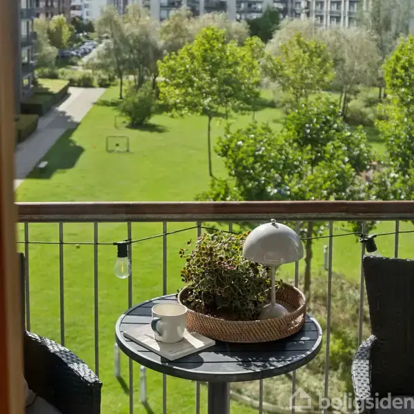 Et bord på en altan rummer en lampe, plante og kop. I baggrunden ses en grøn have med græsplæne og træer i dagslys.