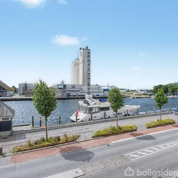 En båd ligger fortøjet ved en kaj langs en vej med træer. I baggrunden ses industrihuse ved vandkanten under en klar blå himmel.