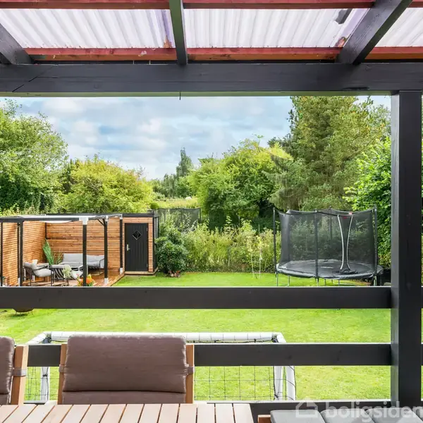 Terrasse med træmøbler, hvor der ses ud over en grøn have med en trampolin og en lille pavillon i hjørnet. Haven er omgivet af buske og træer.