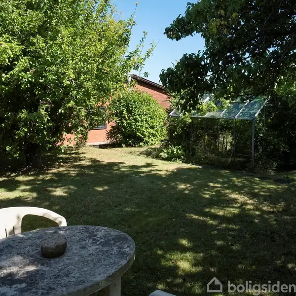 Et rundt stuebord af sten står stille i en solrig have, omgivet af grønne træer og græsplæne. En lille drivhusstruktur ses i baggrunden ved siden af et rødt hus.