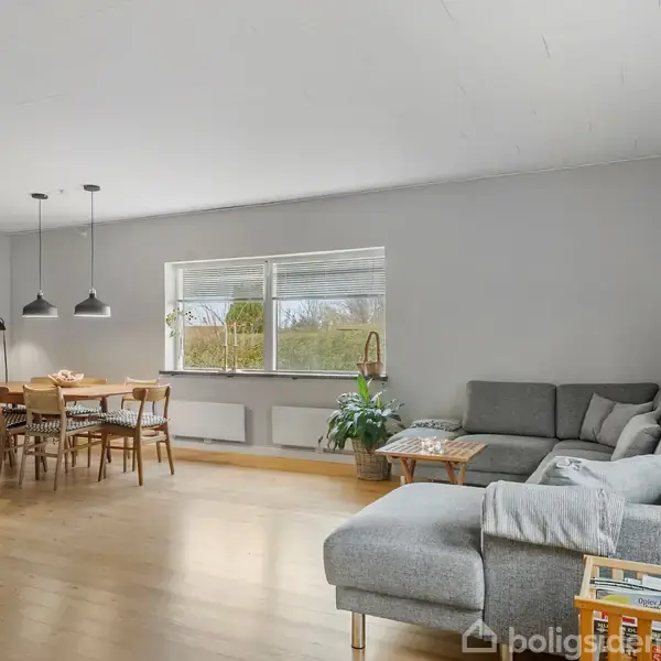 Stor stue med en grå hjørnesofa til højre, et spisebord med træstole til venstre, under tre hængelamper. Rummet har lyse vægge, trægulv og et stort vindue.