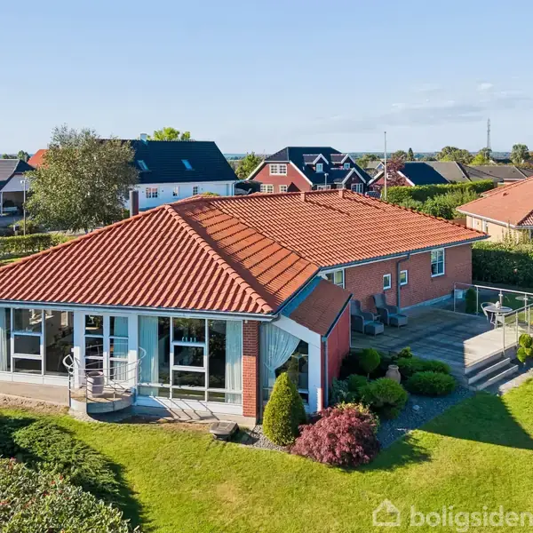En moderne villa med rødt tegltag har store vinduer og en terrasse. Den er omgivet af velplejede græsplæner og buske i et boligkvarter med lignende huse i baggrunden.