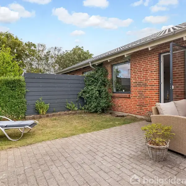 Terrassemøbler står på en brolagt terrasse foran et rødt murstenshus. En liggestol står på græsset ved siden af, omgivet af grønne planter og hække under en klar himmel.