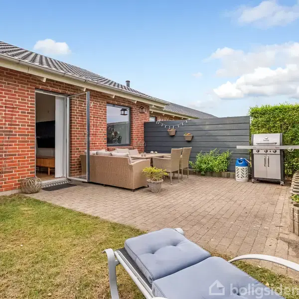 Rødt murstenshus med åben terrassedør til en møbleret terrasse med sofaer og et grillområde. Haven har græsplæne, og hækken skaber en afgrænset, privat atmosfære under en blå himmel.