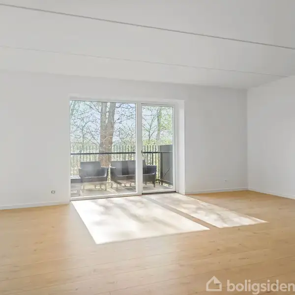 Stue med trægulv og store vinduer, der fører ud til en balkon med stole. Rummet er lyst og tomt, omgivet af hvide vægge.