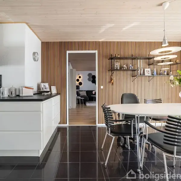 Moderne køkken med hvide skabe og sort emhætte. Spisebord med seks stole i lyst rum. Trævæg med hylder til dekoration. Stor vase med blomster på bordet. Ingen synlig tekst.