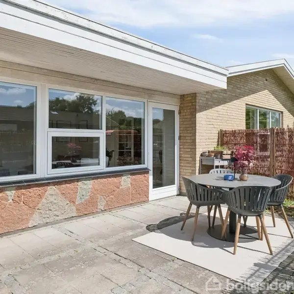 Udendørs terrasse med fire grå stole omkring et rundt bord. Terrassefliser omgiver møblerne. I baggrunden ses en bolig med store vinduer og en dør, omgivet af en murstensfacade.