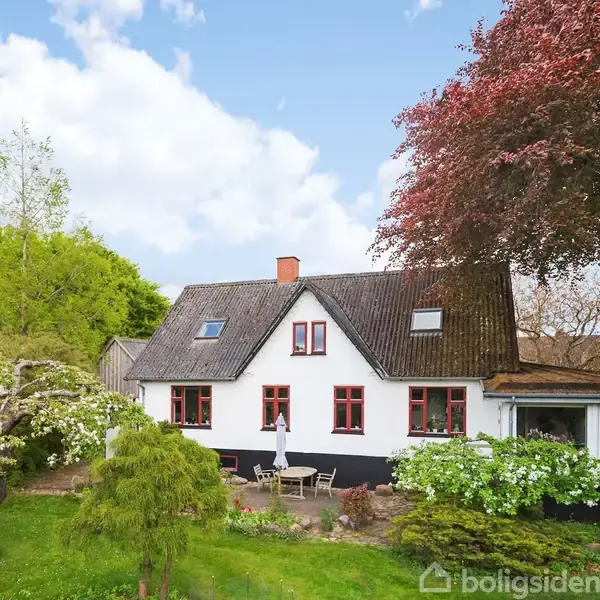 Et hvidt hus med røde vinduer er omgivet af blomstrende træer og buske. En terrasse med borde og stole ligger foran huset under en blå himmel med få skyer.