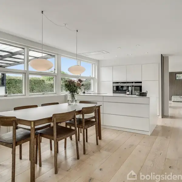 Spisebord med seks stole står ved vinduer. Køkkenø med blomster i vase bagved. Lyse trægulve og moderne indretning. Store vinduer giver udsigt til overdækket terrasse og grønne omgivelser.