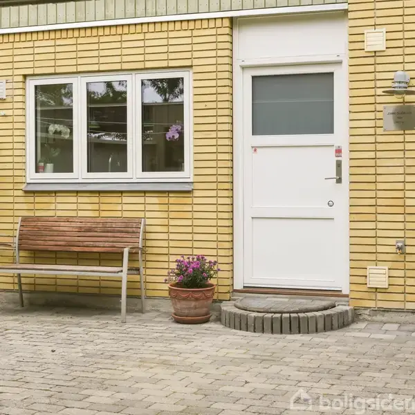 Bænk står foran gul murstensbygning med hvid dør og vindue. En potteplante og cafébord med stole anbringes på flisebelagt terrasse i gårdmiljø.