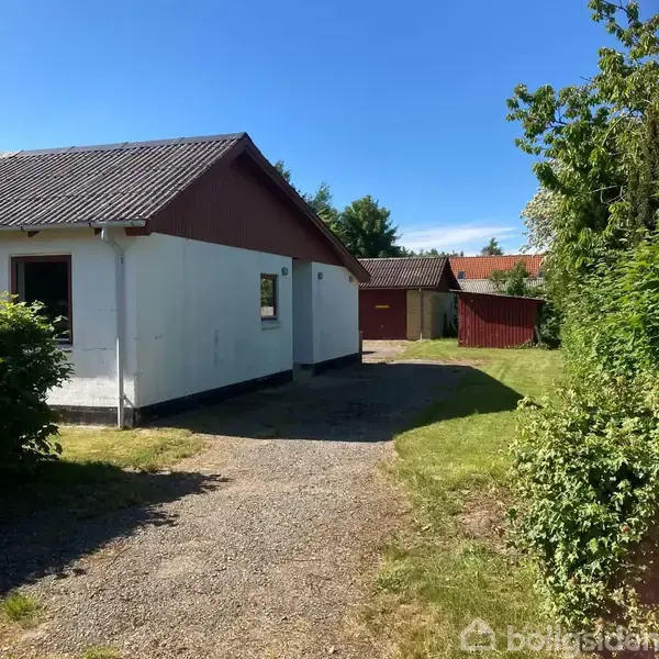 Et hvidt hus med rødt tag står stille ved siden af en grusvej og en grøn have. I baggrunden ses en garage og flere træer.