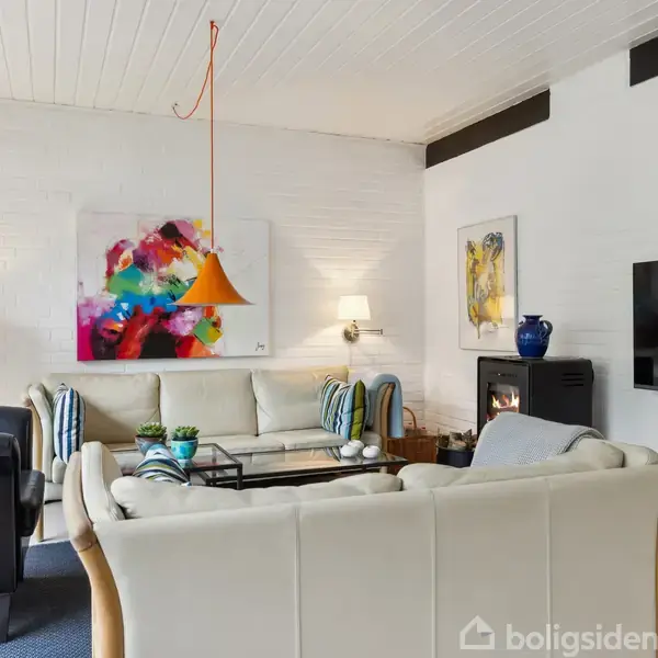 En hyggelig stue med lyse sofaer og en farverig maleri på væggen. Et tv hænger over en pejs. Lamper og pudder tilføjer stemning til rummet.