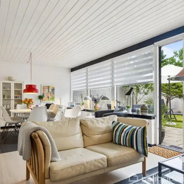 En lys stue med en beige sofa, pyntet med stribede puder, åbner ud mod en have gennem store vinduer. Der er et spisebord med hvide stole og røde lamper.