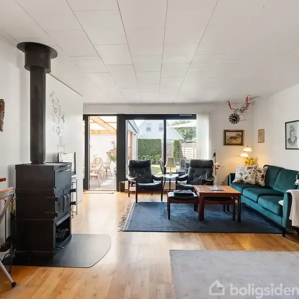 Stue med sort brændeovn ved siden af køkkenet, sofa og lænestole placeret omkring sofabordet. Store vinduer giver udsigt til haven med terrasse. Ingen tekst til stede.