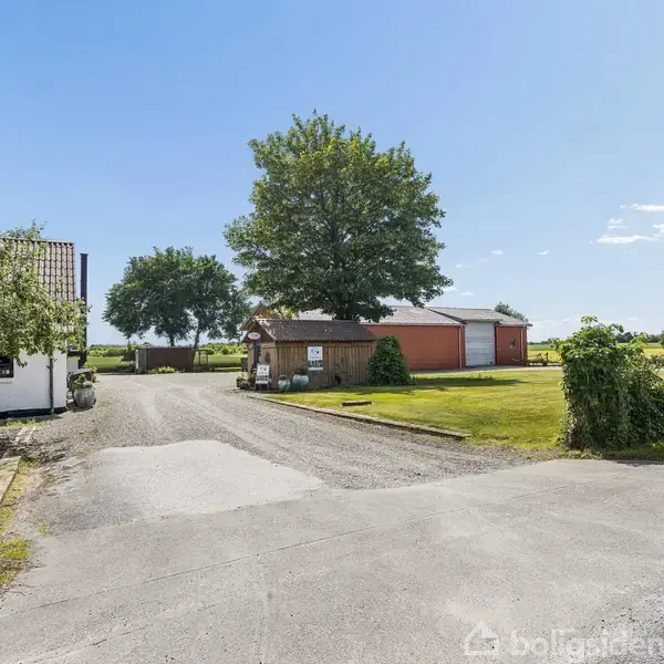 En hvid bygning med et skilt ved indgangen står på en gårdsplads. En stor træstruktur ses i baggrunden omgivet af grønne marker og buske under en klar blå himmel.