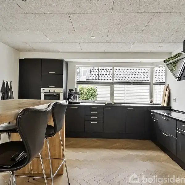 Køkken med sorte skabe og integrerede apparater. En høj træbar med barstole står i forgrunden. Store vinduer giver udsigt til et hus og træer udenfor.