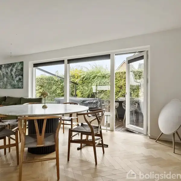 Rundt spisebord omringet af træstole, stående på parketgulv. Store vinduer i baggrunden åbner ud til en frodig have. Et sofahjørne ses til venstre.