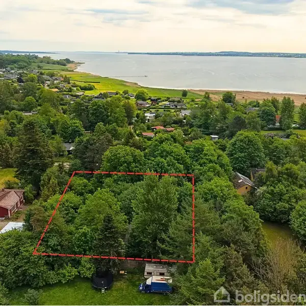 Grønt område markeret med rød streg; omgivet af træer og bebyggelse. I baggrunden ses en kystlinje med hav og sandstrand, der skaber en naturskøn setting.