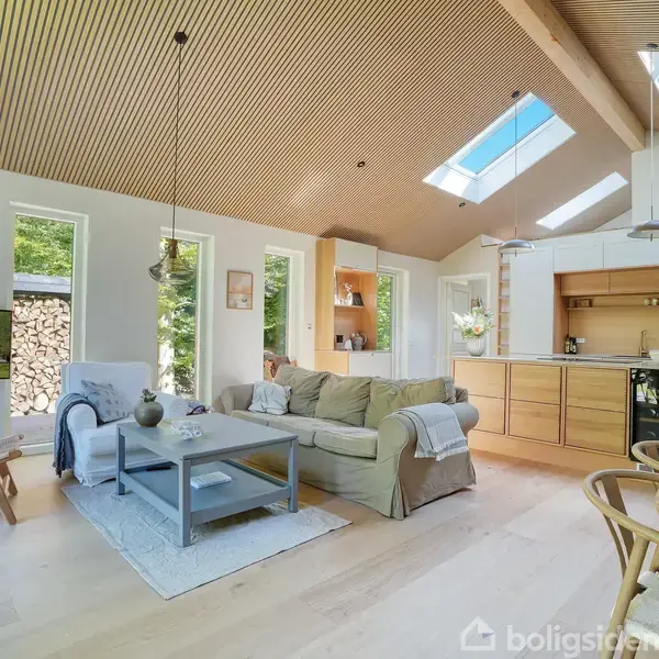 Stue med sofa og sofabord, belyset af ovenlys. Køkken i baggrunden med træskabe. Lyse træpaneler i loftet. Store vinduer giver udsigt til natur udenfor.