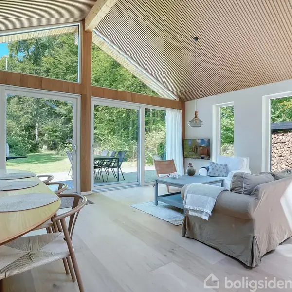 Stue med sofa og bord vendt mod store vinduer, der viser en skovudsigt. Rundt spisebord står til venstre med blomster og dækkeservietter. Lyse trælofter og gulve skaber en lys atmosfære.