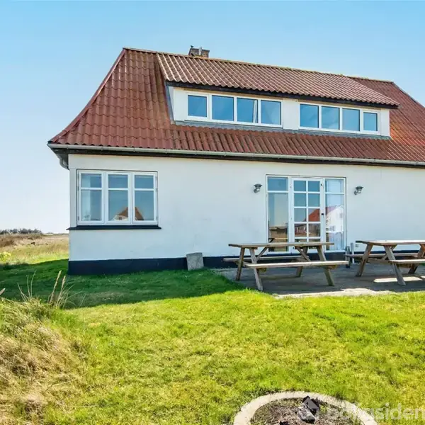 Et hvidt hus med rødt tag står på en grøn græsplæne. To picnicborde er placeret foran huset. I baggrunden ses åbne marker og en klar himmel.