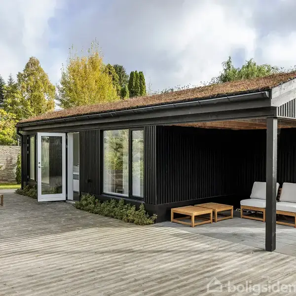 Et sort træhus med åbne dobbeltdøre står på en terrasse med liggestole. Der er grønt tag. Haven er omgivet af træer og buske, skaber en fredelig atmosfære.