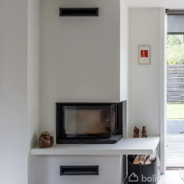 En indbygget pejs med sort glasfront er placeret i en hvid væg. Dekorationer står på hylden, og et lille billede hænger ved siden af. Udenfor ses græsplæne og hegn.