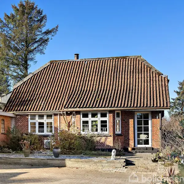 Et rødt murstenshus med skråt tag står i en have. Omkranset af træer, viser huset flere vinduer, en hoveddør og en garage til venstre.