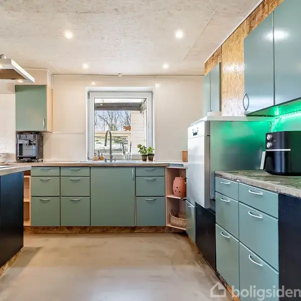 Et moderne køkken med grønne skabe og skuffer er indrettet langs to vægge, med en kaffemaskine og ovn indbygget. I midten ses et vindue med planter udenfor.