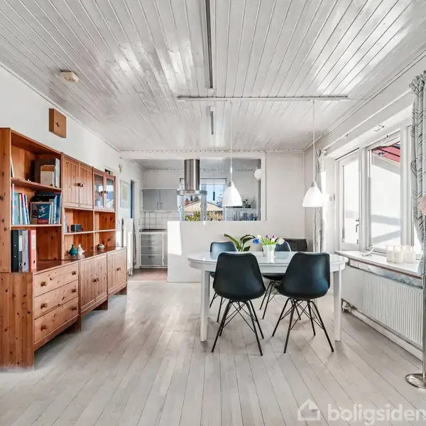 Spisebord med sorte stole står i lyst, åbent rum med hvidmalede trægulve og -lofter. Til venstre er en træreol med bøger, og store vinduer slipper lys ind. lamp om bordet.