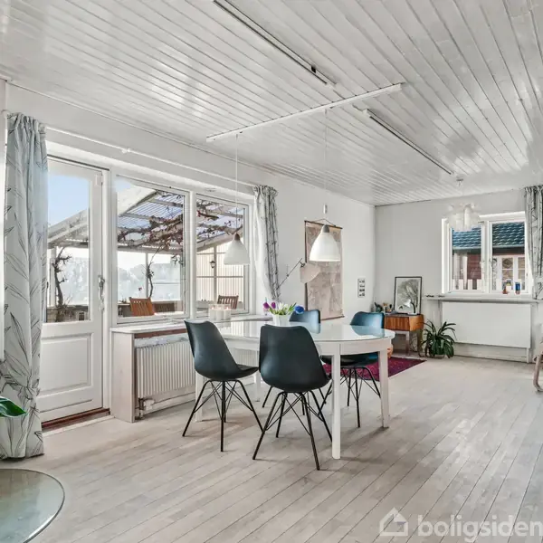 Spisebord med sorte stole står i et lyst rum med trægulv og hvide vægge. Store vinduer med gardiner lader naturligt lys strømme ind. Plante i hjørnet.