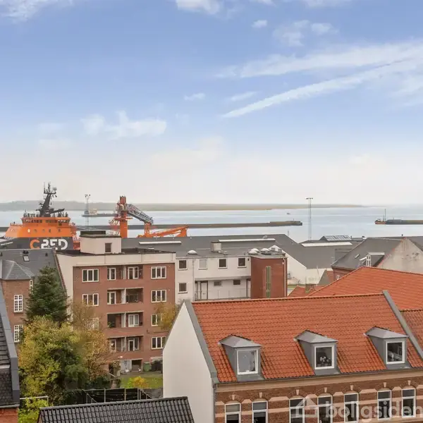 En stor orange båd ligger ved havnen og er omgivet af mange grå og røde bygninger. I baggrunden ses vand og en klar blå himmel med lette skyer.