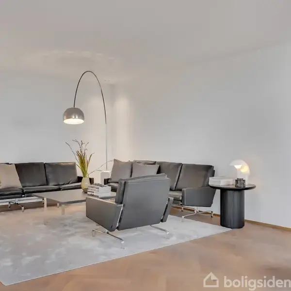 En moderne stue med sorte lædersofaer og en buet gulvlampe, placeret på et lyst tæppe. Et rundt sidebord med lampe og en plante tilføjer hygge til rummet.