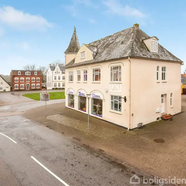 Hvidt to-etagers bygning med skiferbelagt tag står på en hjørnegrund, hvor veje mødes. Bygningen har store vinduer i stueetagen, og der er omkringliggende bygninger og en brostensbelagt vej.