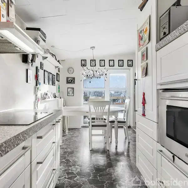 Et lyst køkken med hvide skabe og grå bordplader rummer et spisebord med stole. Der hænger en lysekrone over bordet, og væggen har flere indrammede billeder.