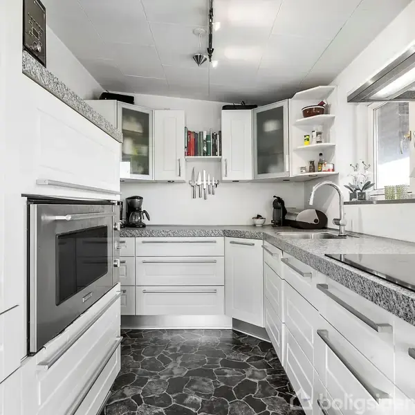 Hvidt moderne køkken med ovn, kogeplade og vask, hvor køkkenmaskiner og kogebøger er placeret på bordplader og hylder. Sorte flisegulve med et vævet Spiderman-ophæng nær ovnen.