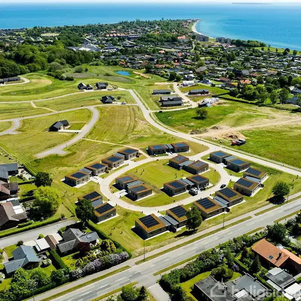 Nybyggeri ses fra luften; huse danner en cirkulær formation. Omkringliggende græsmarker og veje strækker sig mod en by ved kysten i baggrunden. Havet skinner i horisonten.
