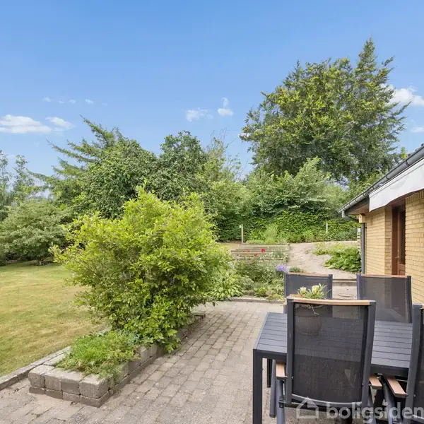 Terrassemøbler står stille på en flisebelagt terrasse ved siden af et gulstenshus. Baggrunden består af en grøn have med græsplæne og tætte buske og træer under en klar blå himmel.